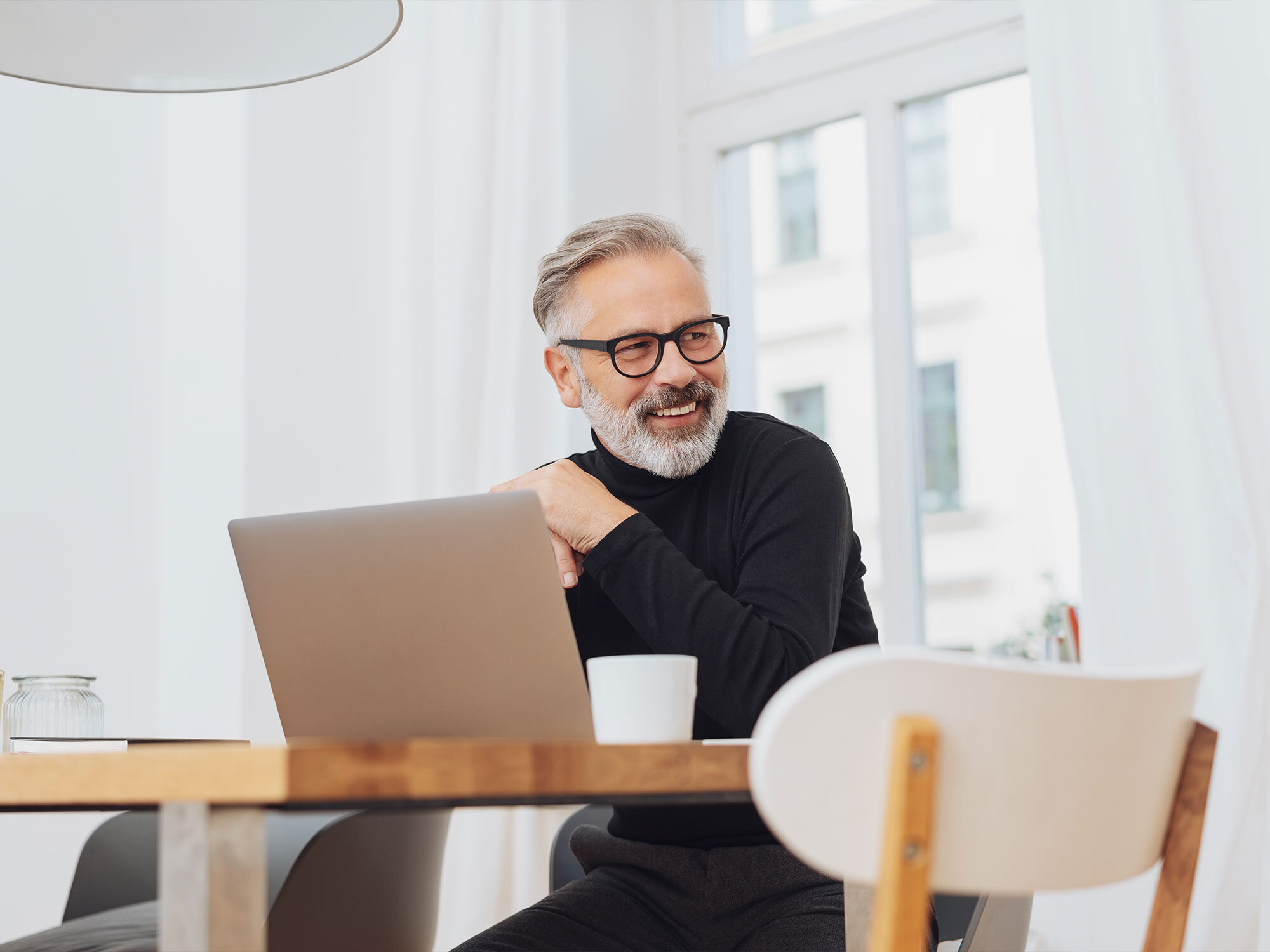 Ein älterer Versicherungsmakler sitzt entspannt zu Hause vorm Laptop und schaut lächelnd zur Seite
