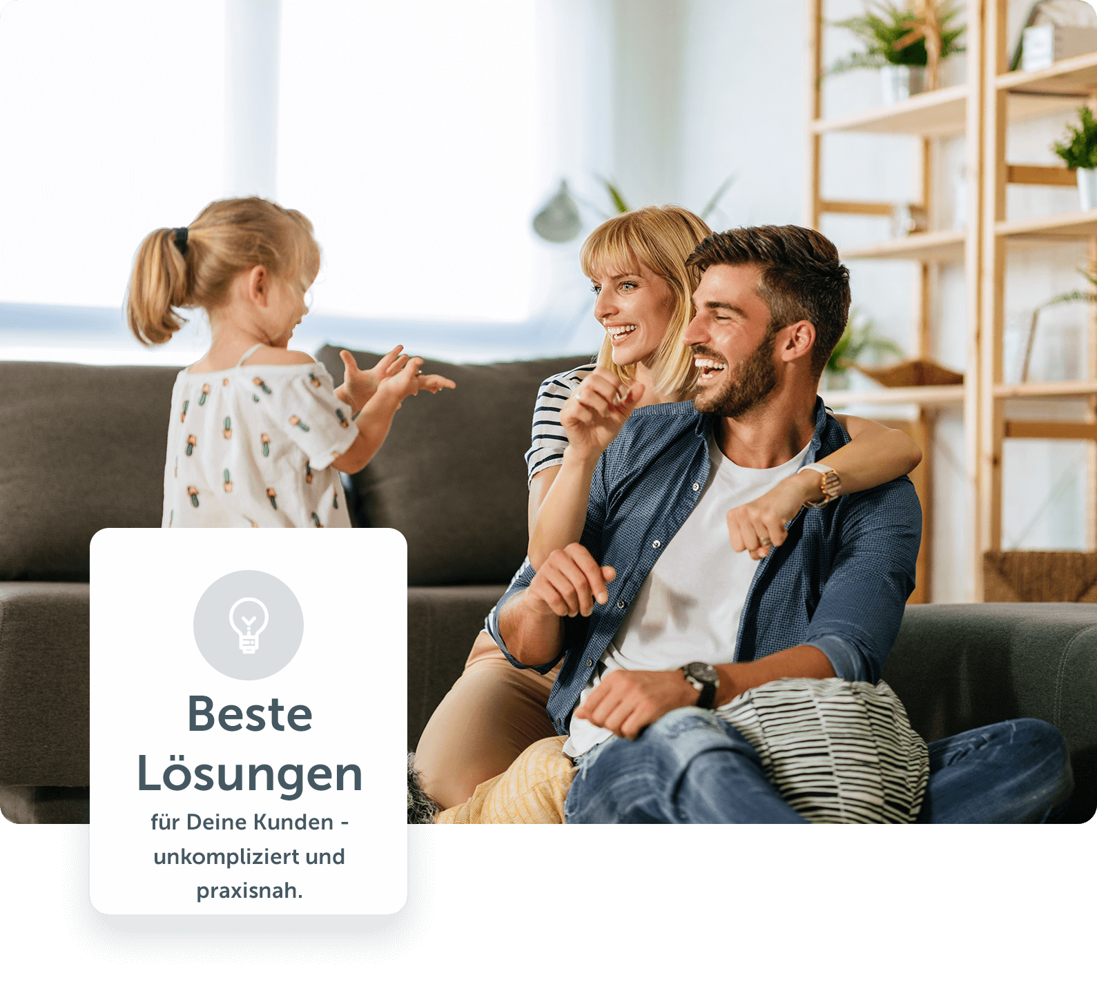 Familie (Vater, Mutter und kleine Tochter) sitzt lachend vor dem Sofa auf dem Boden in einem lichtdurchfluteten Wohnzimmer 