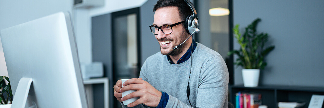 Versicherungsmakler mit Headset und einer Kaffeetasse in der Hand sitzt fröhlich vor dem Bildschirm und nimmt an einer Weiterbildung teil.